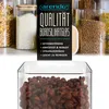 A close-up view of a glass storage container filled with coffee beans, with a marketing overlay detailing product features and a blurred background of other storage containers. Produktbild-Vorschau