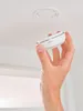 A hand is holding a white smoke detector near a white ceiling mount. Podgląd zdjęcia produktu