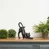 A black cat figurine is displayed on a wooden surface with decorative green spheres and a potted plant in the background. Anteprima dell’immagine prodotto