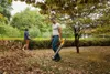 Two people are in a garden, one is using a leaf blower to clear fallen leaves. Produktbild-Vorschau