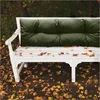 A white garden bench with a dark green tufted cushion, placed outdoors in autumn with fallen leaves scattered on the bench and ground. Náhled obrázku produktu