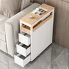 A slim white cabinet with wood accents and three drawers sits next to a sofa, with a cup and magazines on top. Produktbild-Vorschau