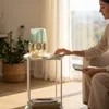 A woman is sitting next to a small side table with a decorative vase and a device on it. Produktbild-Vorschau