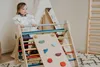 A young child stands on a wooden climbing triangle with a slide attachment in a room with a teepee and a ladder in the background. Produktbild-Vorschau