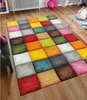 A colorful, modern rug with a checkered design is laid out on a wooden floor in a living room setting. A green armchair is partially visible on the left, and a coffee table with decorative items is on the right. Produktbild-Vorschau