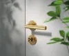 A close-up shot of a gold door handle and keyhole on a white door, with blurred green leaves in the background. Produktbild-Vorschau