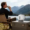 A man is sitting in a camping chair by a lake, drinking from a mug, with a small electric kettle on a cooler next to him. Mountains and trees are in the background. Produktbild-Vorschau