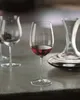 A close-up shot of a wine glass filled with red wine, with another empty wine glass and a decanter in the background, all placed on a marble countertop. Produktbild-Vorschau