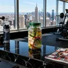 Glas-Salatglas mit grünem Deckel, gefüllt mit frischem Salat, neben einer Wasserflasche auf schwarzer Marmorarbeitsplatte in einem Fitnessstudio mit Stadtblick Podgląd zdjęcia produktu
