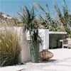 A green planter with a plant in it sits on a patio next to a woven lantern. In the background, there is a hot tub and tall grasses. Voir les images du produit