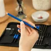 A hand holding a blue pencil with a pencil case in the background. Podgląd zdjęcia produktu