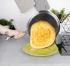 A hand is holding a frying pan with a pancake in it, tilting it over a green plate. Voir les images du produit