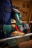 A person wearing work clothes and gloves is using a cordless angle grinder to cut a metal pipe in a workshop setting. Sparks are flying from the cut. Produktbild-Vorschau