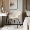 A cream-colored armchair with diamond quilting and black metal legs is placed next to a white vanity table with a mirror and some cosmetic products. The scene is set in a bedroom with a bed visible on the right. Produktbild-Vorschau