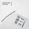 A close-up of a white fitted sheet, showing its texture and elasticized edge, with marketing text and care symbols. Produktbild-Vorschau