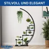 A black metal plant stand with multiple tiers, holding various potted plants, with hanging plants above. The background is a plain wall with a framed picture on the right. Produktbild-Vorschau
