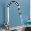 A stainless steel kitchen faucet with a pull-out spray head is shown in use, with water flowing into a sink. A glass of blue liquid is visible to the right. Produktbild-Vorschau