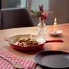 A cast iron pan with food is placed on a wooden table, next to a candle and a vase of flowers. Produktbild-Vorschau