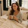 A woman is sitting in bed, wrapped in a blanket, holding a mug. Anteprima dell’immagine prodotto