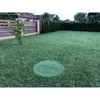 A green manhole cover sits on a lush green lawn in front of a wooden fence and a row of evergreen trees. Podgląd zdjęcia produktu