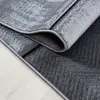 A close-up view of a rolled-up rug with a modern abstract pattern in shades of gray and black, placed on a light wooden floor. Voir les images du produit