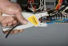 A person's hand is using a wire stripper tool on a black cable, with electrical wiring and components in the background. Anteprima dell’immagine prodotto