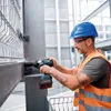 A construction worker wearing a blue hard hat and an orange safety vest is using a cordless impact wrench to tighten a bolt on a metal structure. Podgląd zdjęcia produktu