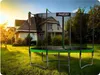 A trampoline is set up in a grassy backyard with a house in the background during sunset. Produktbild-Vorschau