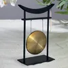 A decorative gong with a black stand and mallets, set against a light, textured background with hints of greenery and white flowers. Náhľad obrázku produktu