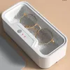 A close-up of a white ultrasonic cleaner with gold-rimmed glasses inside, sitting on a tan surface. Produktbild-Vorschau