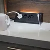 A black power strip with three outlets and a USB-C port is shown on a white countertop, with a white mug to the left and a wooden backsplash behind it. An orange light emanates from the top of the power strip. Produktbild-Vorschau