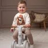A young child is sitting on a balance bike indoors, with a chair and pillow in the background. Náhľad obrázku produktu