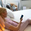 A woman in pajamas is lying on a bed and looking at her phone. Produktbild-Vorschau