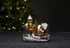 A miniature Christmas village scene with a church and houses, set against a blurred Christmas tree background. Podgląd zdjęcia produktu