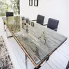 A dining table with a glass top and a wooden base, set in a bright room with chairs. Produktbild-Vorschau