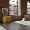 An interior shot of a room with a brick wall, large windows, and sunlight streaming in. A wooden cube stand with a framed artwork is in the foreground, and a leather sofa is visible in the background. Produktbild-Vorschau