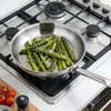Asparagus being cooked in a frying pan on a gas stove, with tomatoes and a pepper grinder visible in the foreground. Náhled obrázku produktu
