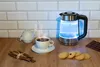 A glass electric kettle with blue LED lighting is shown on a wooden countertop next to a cup of tea, a small teapot, cinnamon sticks, and cookies. Náhled obrázku produktu