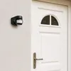 A black motion detector is mounted on a textured white wall next to a white door with a silver handle. Náhľad obrázku produktu