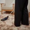 A person is vacuuming a fluffy beige rug in a room with a wooden chair and white curtains. Náhled obrázku produktu