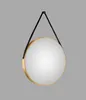 A round mirror with a gold frame and a black leather-look strap hanging against a plain gray background. Náhled obrázku produktu