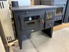 A dark gray wood-burning stove with a glass door and oven is displayed in a warehouse or showroom setting. Anteprima dell’immagine prodotto
