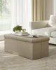 A beige storage ottoman is placed on a textured rug in a living room. Books and a plant are on top of the ottoman, with a couch visible in the background. Náhled obrázku produktu
