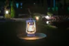 A white kerosene lantern is lit and placed on a dark circular surface in a garden at night. Voir les images du produit