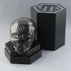 A faceted skull-shaped object, likely a perfume bottle, is displayed on a black stand next to its hexagonal black packaging box, which features a silver logo. Produktbild-Vorschau