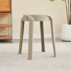 A beige stool is placed on a textured rug in a room with a wooden shelf unit and a potted plant in the background. Voir les images du produit