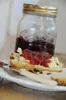 A scone with jam and butter is on a plate, with a jar of jam in the background. Voir les images du produit