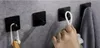 A hand is shown hanging a towel on a black adhesive hook mounted on a gray tiled wall. Other hooks with items are visible. Anteprima dell’immagine prodotto