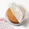A round cake on a white plate, partially covered by parchment paper. A red and white checkered cloth is visible on the left. Produktbild-Vorschau