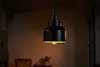 A black pendant light fixture hangs in a dimly lit room, with a glimpse of furniture in the background. Produktbild-Vorschau